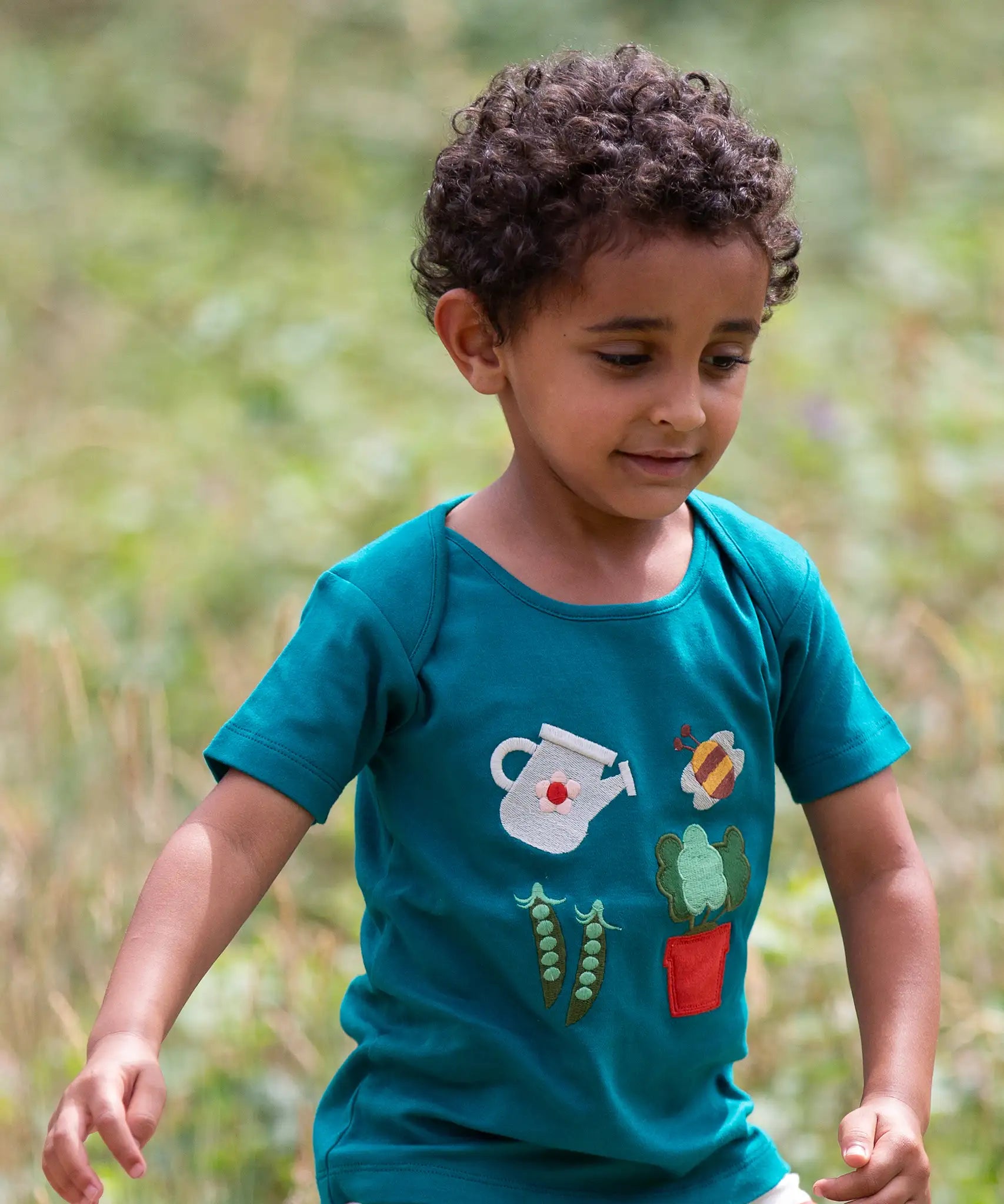 Child wearing the Little Green Radicals envelope neck Spring garden embroidered organic cotton green t-shirt. Features 4 embroidered elements, a white watering can, peas in their pods, a bee and a potted plant.