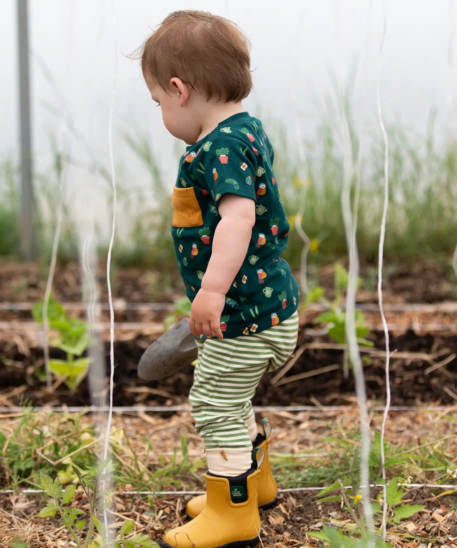 Child wearing the Little Green Radicals Spring garden organic cotton playset outfit with yellow gold wellingtons. Repeat pattern green t-shirt and green and cream striped joggers. 