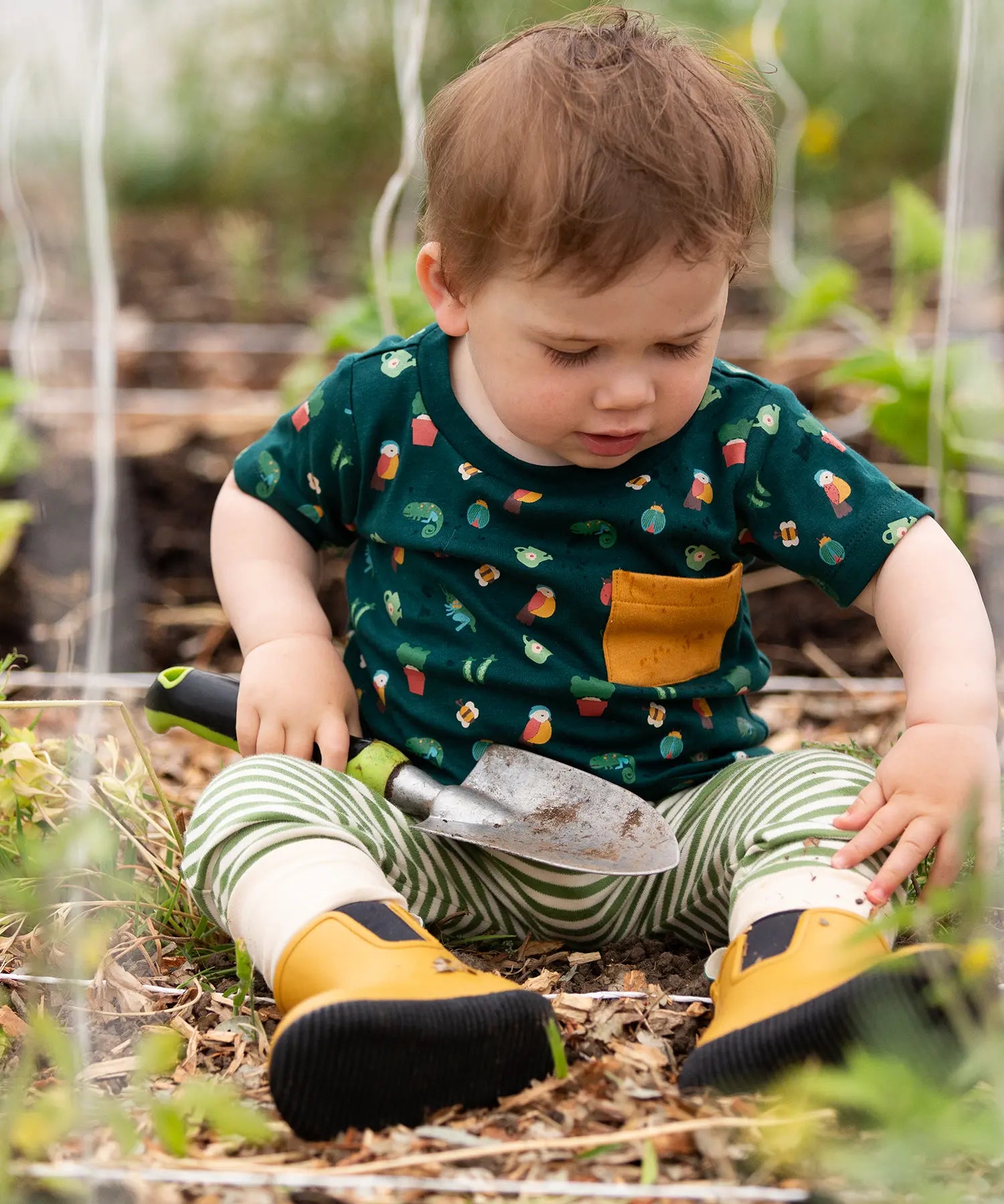 Child wearing the Little Green Radicals Spring garden organic cotton playset outfit with repeat pattern green t-shirt and green and cream striped joggers with yellow wellingtons. 