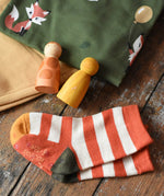 Orange and cream striped socks from the Little Green Radicals mushroom organic cotton socks 5 Pack placed on a wooden surface next to Maxomorra clothing and Grapat peg dolls.