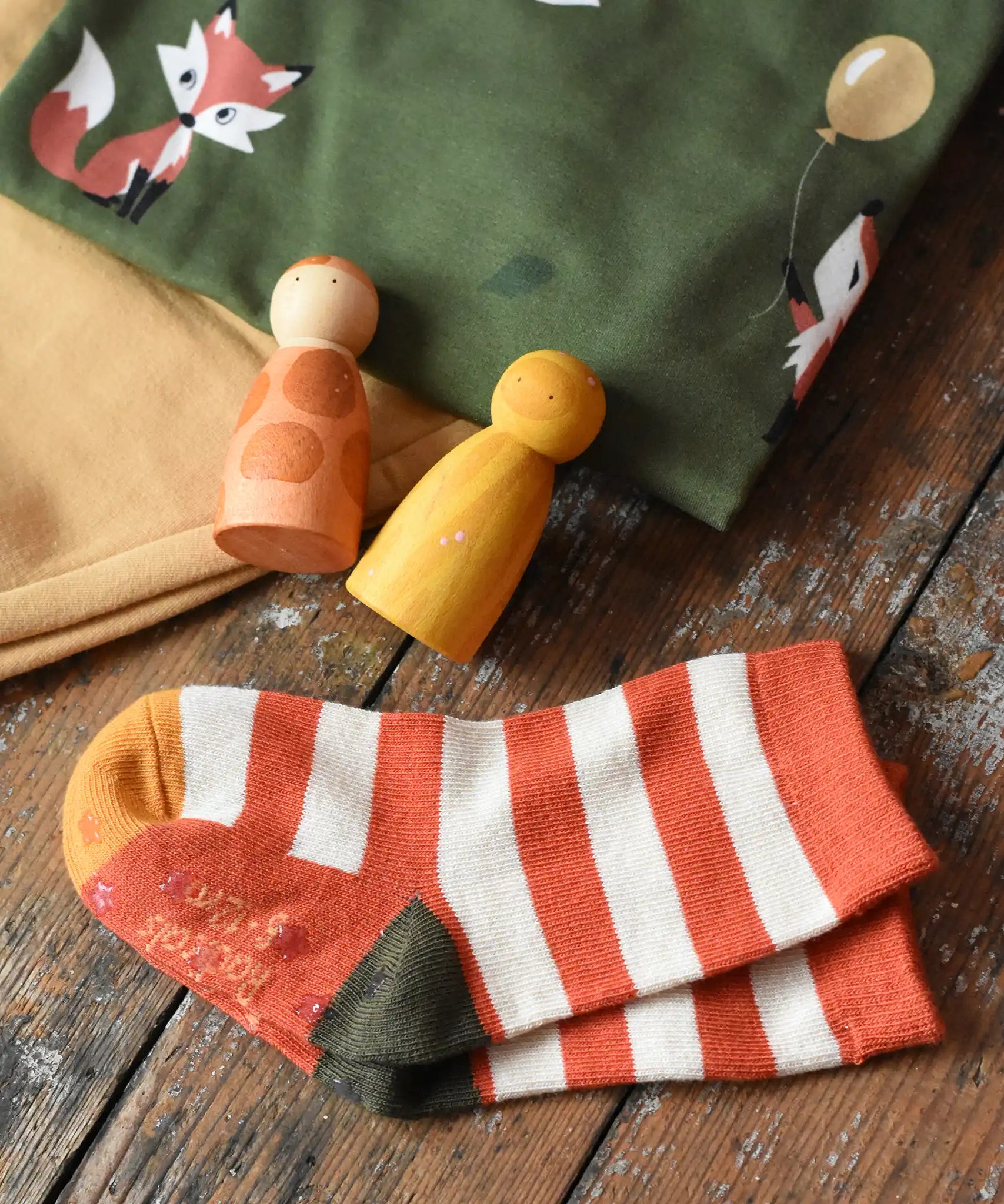 Orange and cream striped socks from the Little Green Radicals mushroom organic cotton socks 5 Pack placed on a wooden surface next to Maxomorra clothing and Grapat peg dolls.