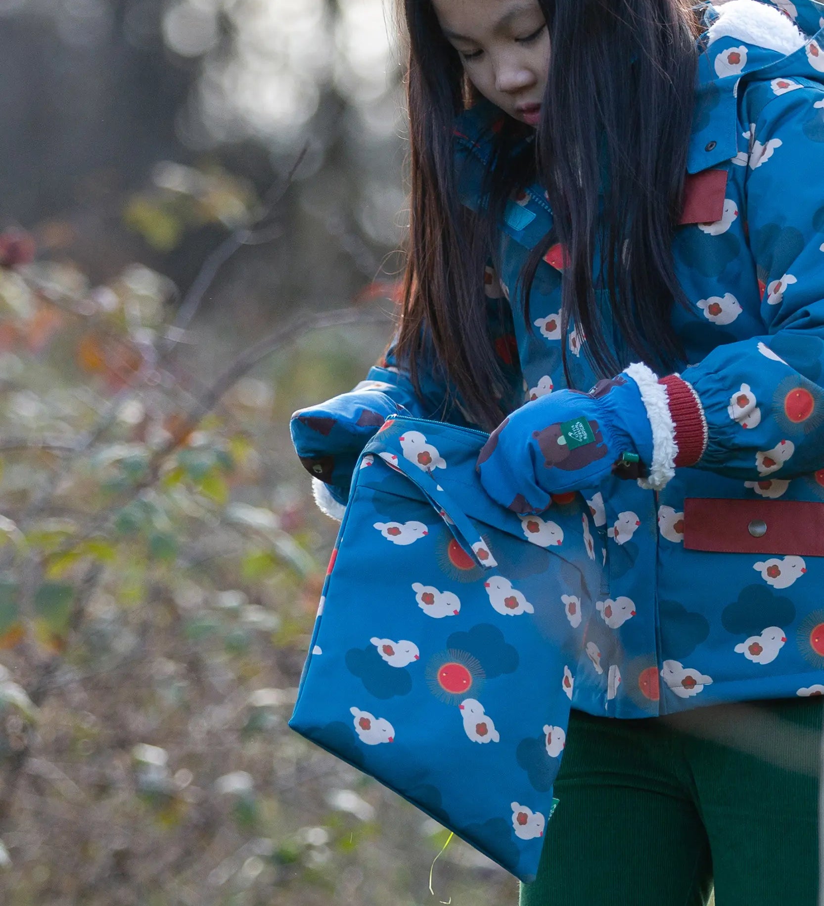 Child holding a Little Green Radicals  sun birds print blue recycled waterproof wet bag wearing a coat and gloves in the same print.