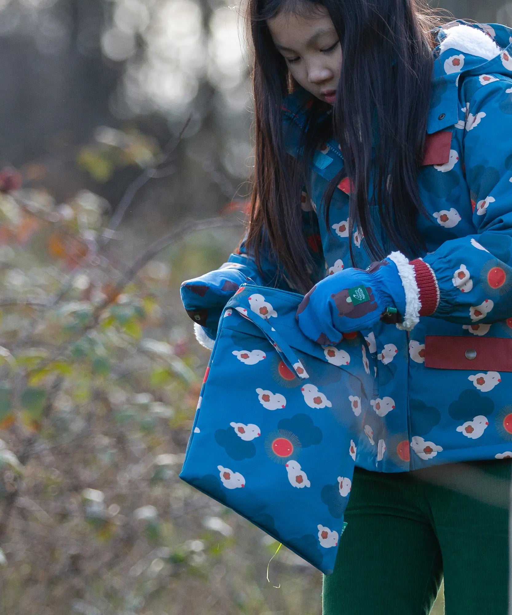 Child holding a Little Green Radicals  sun birds print blue recycled waterproof wet bag wearing a coat and gloves in the same print.