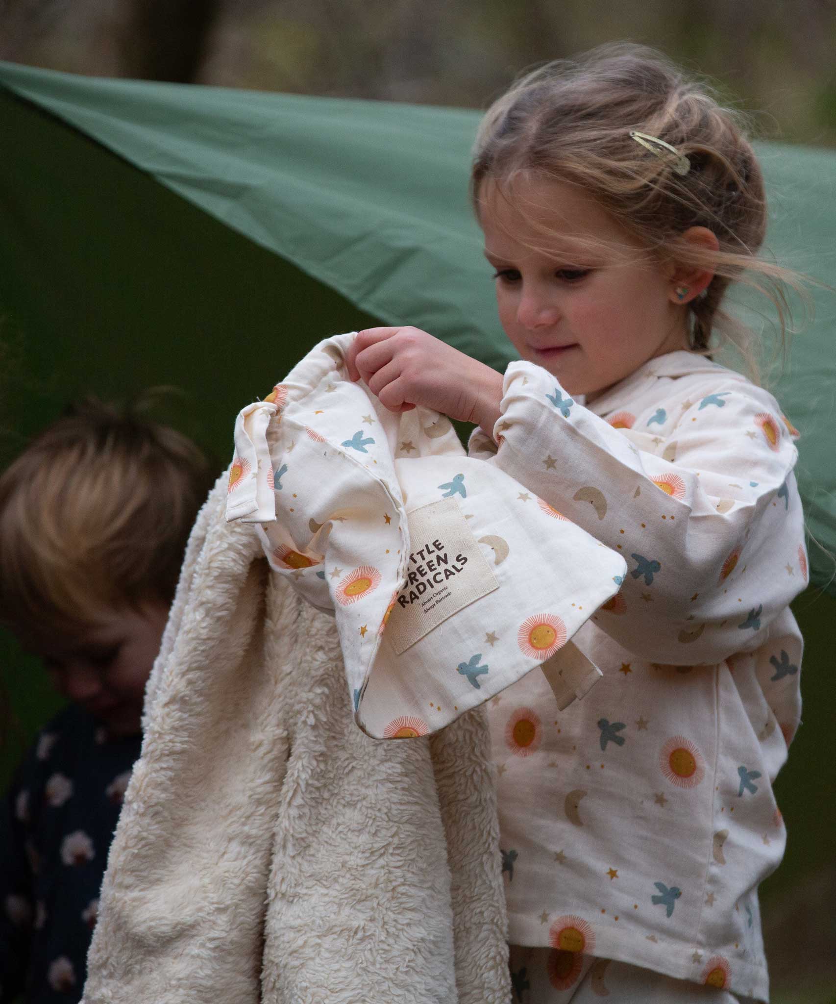 A child holding up the Little Green Radicals moon and sun print Organic Sherpa Hooded Baby Blanket in front of them showing off the fleecy side to the camera. The child is wearing matching pyjamas. This blanket is a part of a wide range of organic cotton clothing and accessories for children available here at Babipur. 

