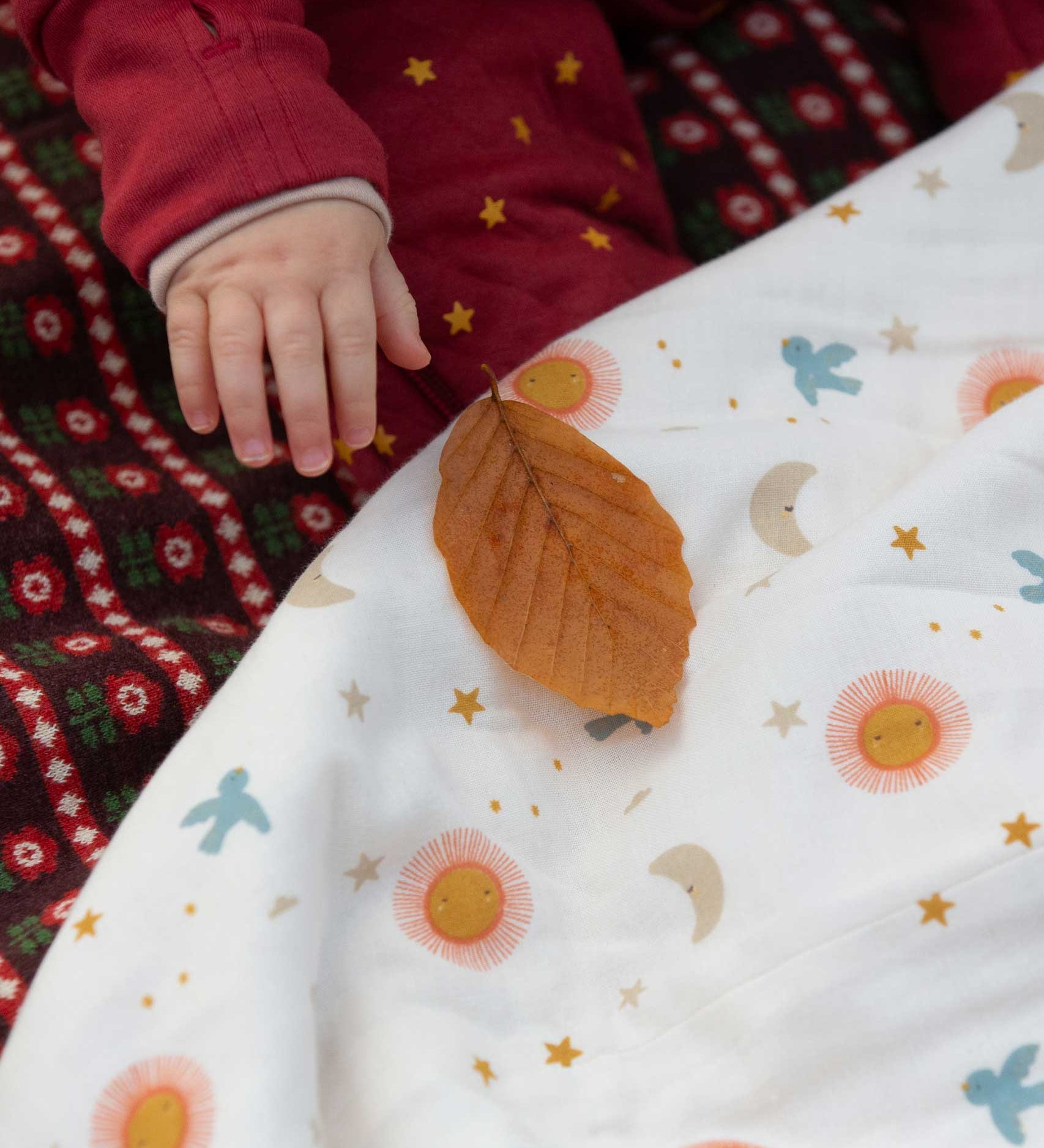 A close up of a babies hand reaching for a leaf that has been placed on the Little Green Radicals moin and sun print Organic Sherpa Hooded Baby Blanket in front of them. This blanket  is a part of a wide range of organic cotton clothing and accessories for children available here at Babipur. 
