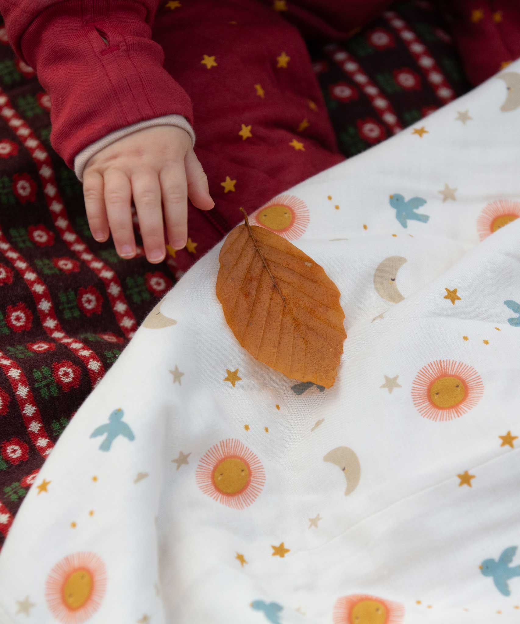 A close up of a babies hand reaching for a leaf that has been placed on the Little Green Radicals moin and sun print Organic Sherpa Hooded Baby Blanket in front of them. This blanket  is a part of a wide range of organic cotton clothing and accessories for children available here at Babipur. 
