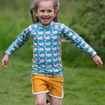 A child wearing the Little Green Radicals Children's Blue Whales design UPF 50+ Sunsafe Long Sleeve Rash Vest with yellow gold swim shorts.