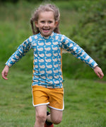 A child wearing the Little Green Radicals Children's Blue Whales design UPF 50+ Sunsafe Long Sleeve Rash Vest with yellow gold swim shorts.