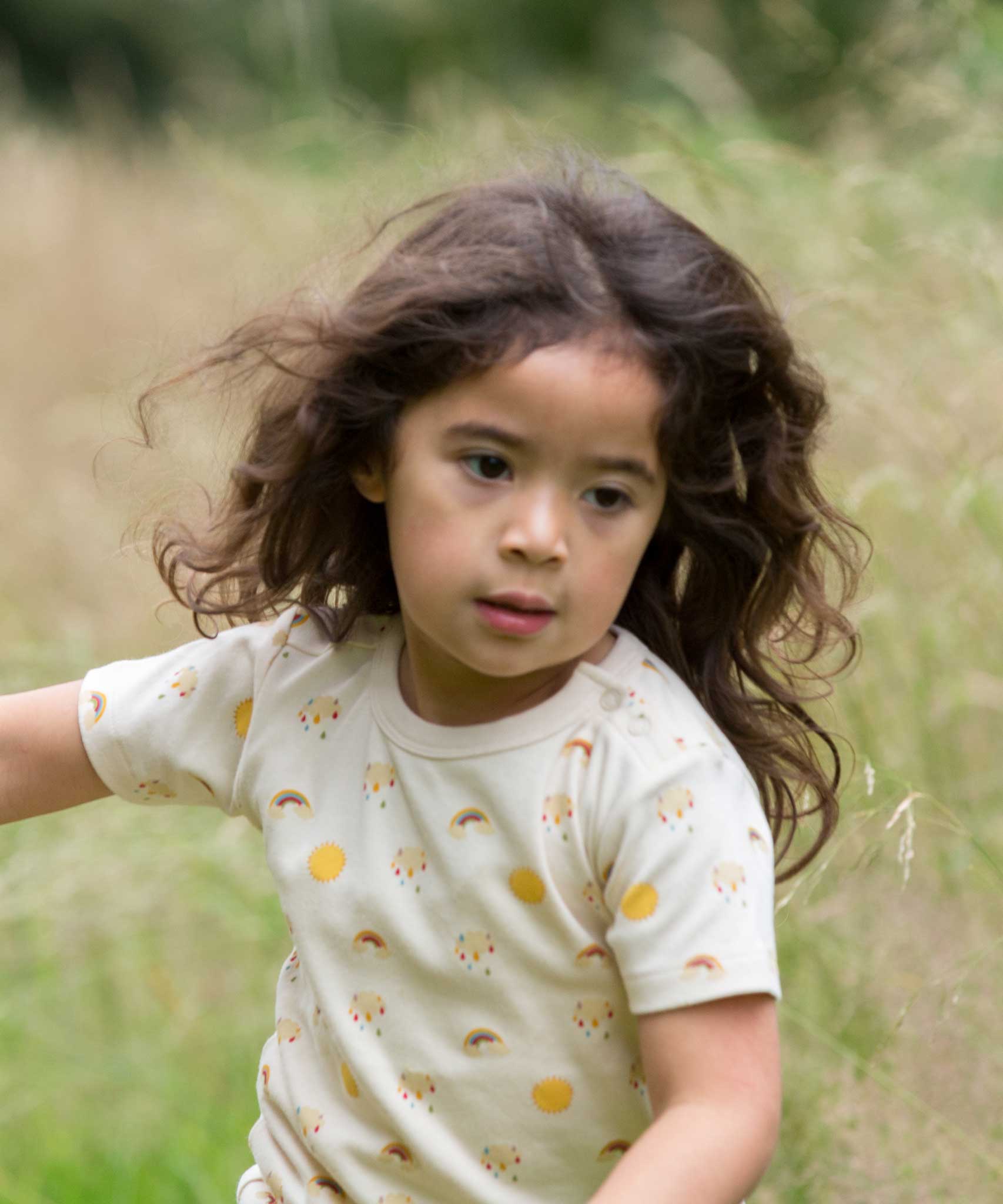 A child running, wearing the Little Green Radicals Sunshine & Rainbows Short Sleeve organic cotton T-Shirt. An off white, cream coloured t-shirt with an all over print featuring rainbows, sunshine and rainbow rainclouds. Little green Radicals have a wide range of organic cotton, Fairtrade children's clothing and outerwear available here at Babipur.