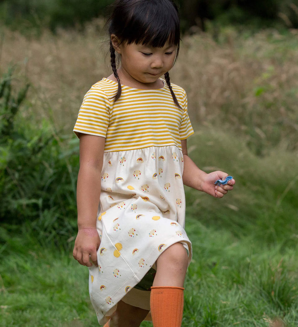 Close up of girl walking in a grass field wearing the Little Green Radicals organic cotton sunshine and rainbows easy peasy dress