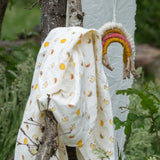 The rainows and sunshine peint muslin cloth placed on a stick outdoors. A Little Green Radicals Sunshine & Rainbows Organic Baby Muslin Set. A  2 Pack of muslin cloths, one is plain yellow gold and the other has an all over print featuring rainbows, sun and rainbow rain clouds. Little green Radicals have a wide range of organic cotton, Fairtrade children's clothing and outerwear available here at Babipur.