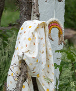 The rainows and sunshine peint muslin cloth placed on a stick outdoors. A Little Green Radicals Sunshine & Rainbows Organic Baby Muslin Set. A  2 Pack of muslin cloths, one is plain yellow gold and the other has an all over print featuring rainbows, sun and rainbow rain clouds. Little green Radicals have a wide range of organic cotton, Fairtrade children's clothing and outerwear available here at Babipur.