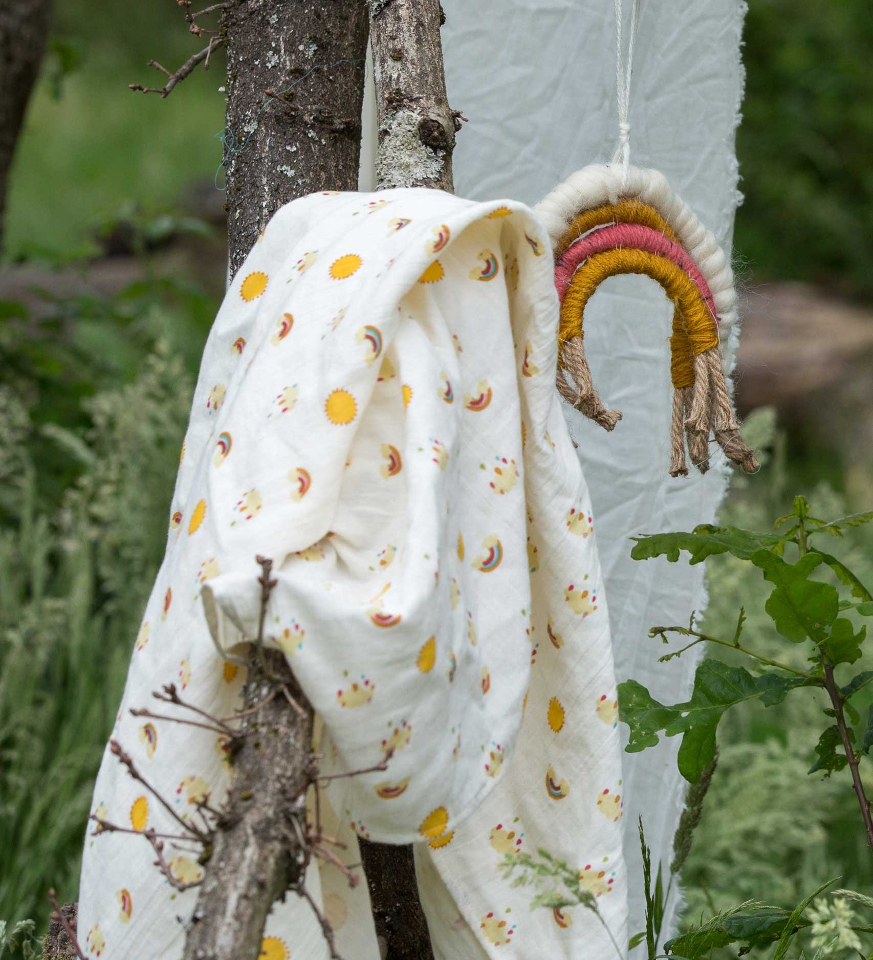 The rainows and sunshine peint muslin cloth placed on a stick outdoors. A Little Green Radicals Sunshine & Rainbows Organic Baby Muslin Set. A  2 Pack of muslin cloths, one is plain yellow gold and the other has an all over print featuring rainbows, sun and rainbow rain clouds. Little green Radicals have a wide range of organic cotton, Fairtrade children's clothing and outerwear available here at Babipur.