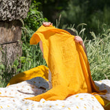 A child holding up a yellow gold coloured muslin cloth in front of them. A Little Green Radicals Sunshine & Rainbows Organic Baby Muslin Set. A  2 Pack of muslin cloths, one is plain yellow gold and the other has an all over print featuring rainbows, sun and rainbow rain clouds. Little green Radicals have a wide range of organic cotton, Fairtrade children's clothing and outerwear available here at Babipur.