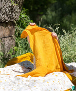 A child holding up a yellow gold coloured muslin cloth in front of them. A Little Green Radicals Sunshine & Rainbows Organic Baby Muslin Set. A  2 Pack of muslin cloths, one is plain yellow gold and the other has an all over print featuring rainbows, sun and rainbow rain clouds. Little green Radicals have a wide range of organic cotton, Fairtrade children's clothing and outerwear available here at Babipur.