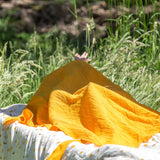 A yellow gold coloured muslin cloth from the Little Green Radicals Sunshine & Rainbows Organic Baby Muslin Set placed on a blanket outdoors. A  2 Pack of muslin cloths, one is plain yellow gold and the other has an all over print featuring rainbows, sun and rainbow rain clouds. Little green Radicals have a wide range of organic cotton, Fairtrade children's clothing and outerwear available here at Babipur.