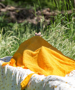 A yellow gold coloured muslin cloth from the Little Green Radicals Sunshine & Rainbows Organic Baby Muslin Set placed on a blanket outdoors. A  2 Pack of muslin cloths, one is plain yellow gold and the other has an all over print featuring rainbows, sun and rainbow rain clouds. Little green Radicals have a wide range of organic cotton, Fairtrade children's clothing and outerwear available here at Babipur.