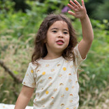 A child waving, wearing the Little Green Radicals Sunshine & Rainbows Short Sleeve organic cotton T-Shirt. An off white, cream coloured t-shirt with an all over print featuring rainbows, sunshine and rainbow rainclouds. Little green Radicals have a wide range of organic cotton, Fairtrade children's clothing and outerwear available here at Babipur.