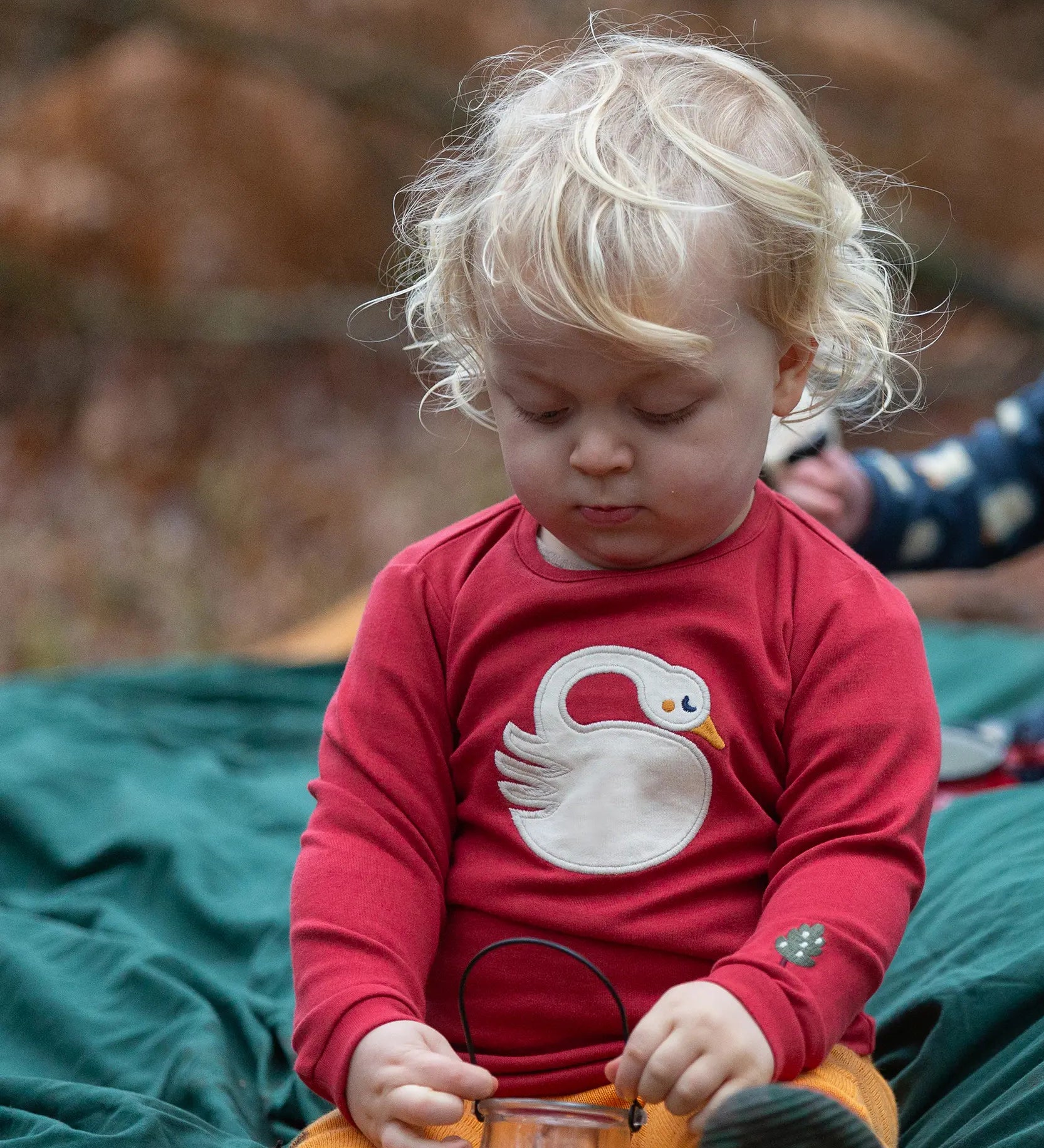 Child wearing the Little Green Radicals sleeping swan applique long sleeve organic cotton top in red with yellow gold joggers sitting on a dark green blanket outdoors.