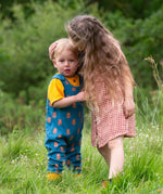 Two children standing in grass,the younger child is wearing the Little Green Radicals Everyday Dungarees with a Teddy Bears print, the older child is wearing a red and white gingham dress.