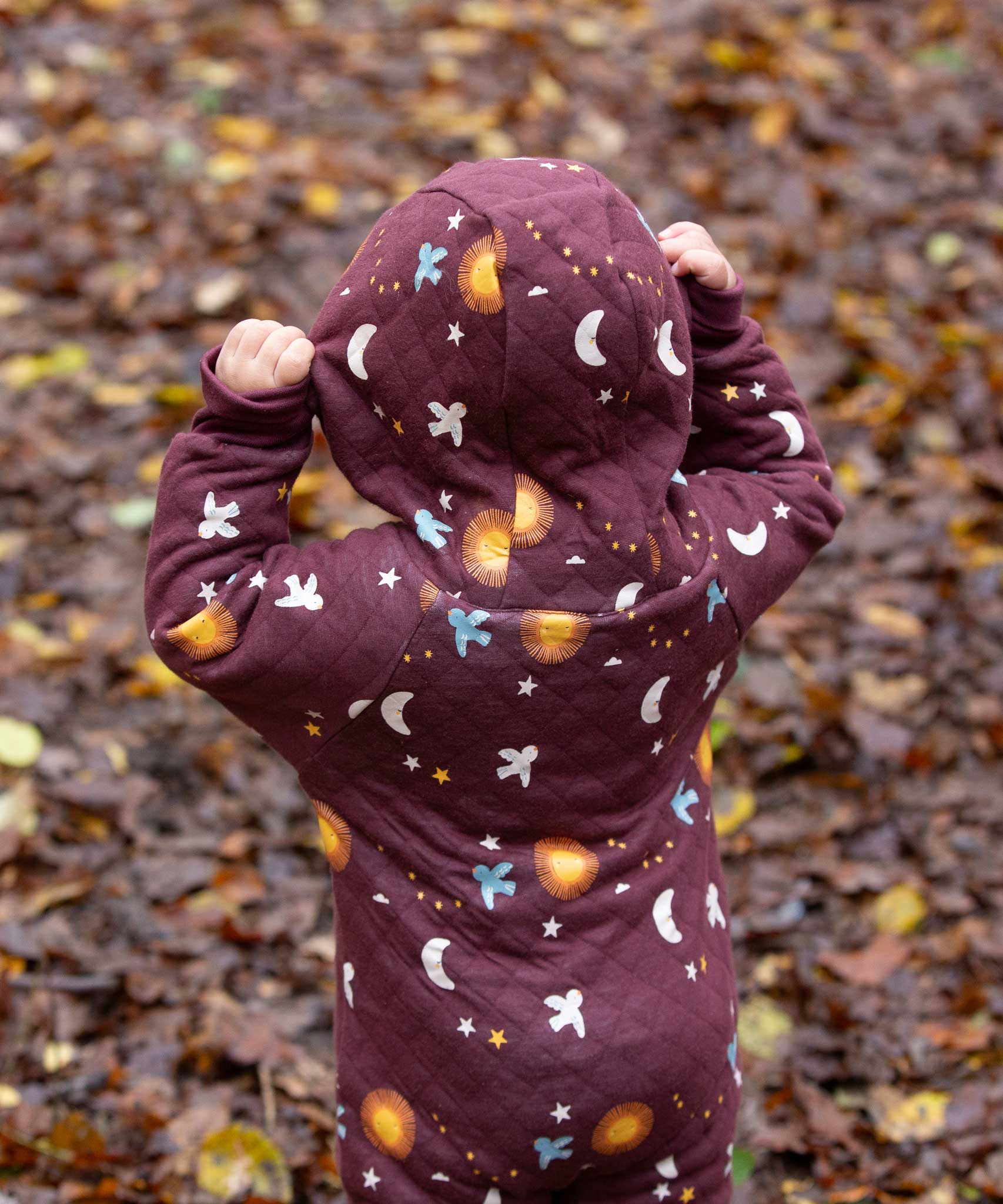 A child wearing the Little Green Radicals Organic Cotton Flying Birds Quilted Snug Suit. The child is facing away from the camera and is wearing the hood up.
A maroon brown coloured suit with an all over print featuring flying birds, stars, moon and  sun. This snug suit is a part of a wide range of organic cotton clothing for children available here at Babipur. 