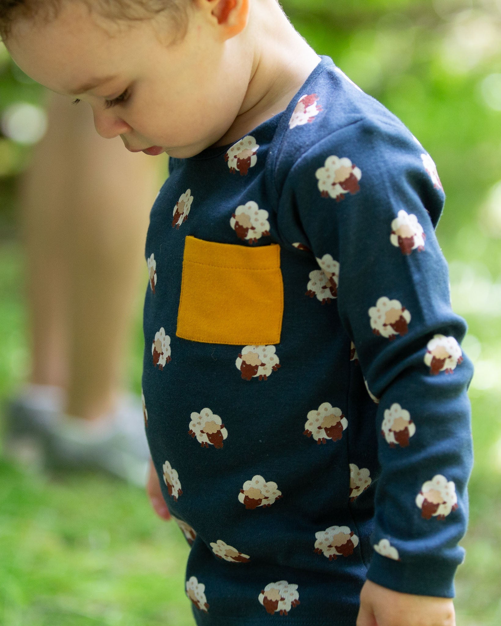 A close up of a child wearing the Little Green Radicals Kids' Organic Top and Joggers Playset with sleeping sheep print. The top has a yellow gold coloured chest pocket.  Little green Radicals have a wide range of organic cotton, Fairtrade children's clothing an outerwear available here at Babipur.