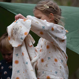 A child wearing the Little Green Radicals moon and sun classic pyjama set and holding up a matching Sherpa lined hooded blanket. A GOTS organic cotton pyjama set including a long sleeve shirt style top in light pink with prints of cute birds flying through stars the moon and sun also includes matching bottoms. These pyjamas are part of a wide range of organic cotton clothing for children available here at Babipur.