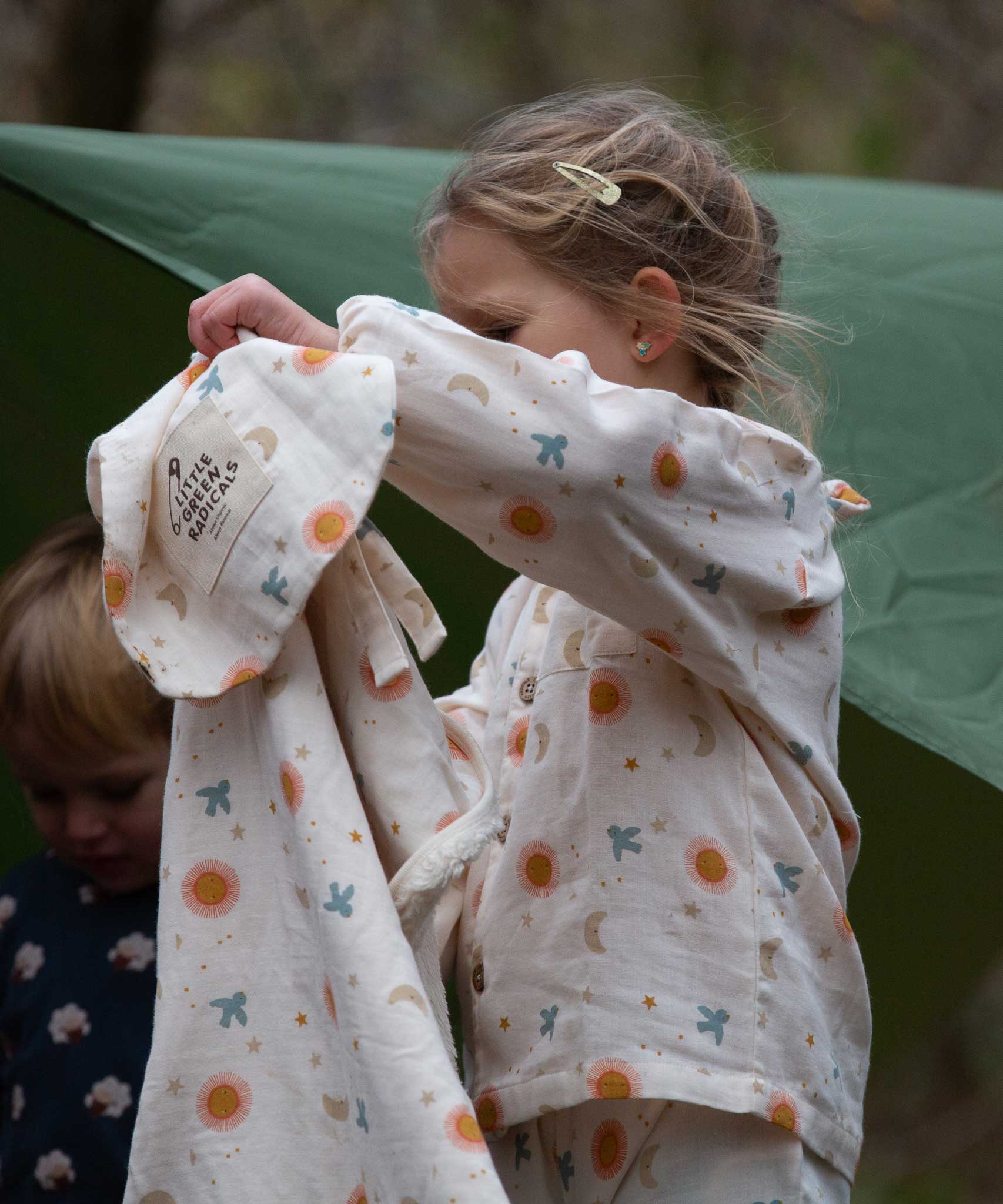 A child wearing the Little Green Radicals moon and sun classic pyjama set and holding up a matching Sherpa lined hooded blanket. A GOTS organic cotton pyjama set including a long sleeve shirt style top in light pink with prints of cute birds flying through stars the moon and sun also includes matching bottoms. These pyjamas are part of a wide range of organic cotton clothing for children available here at Babipur.