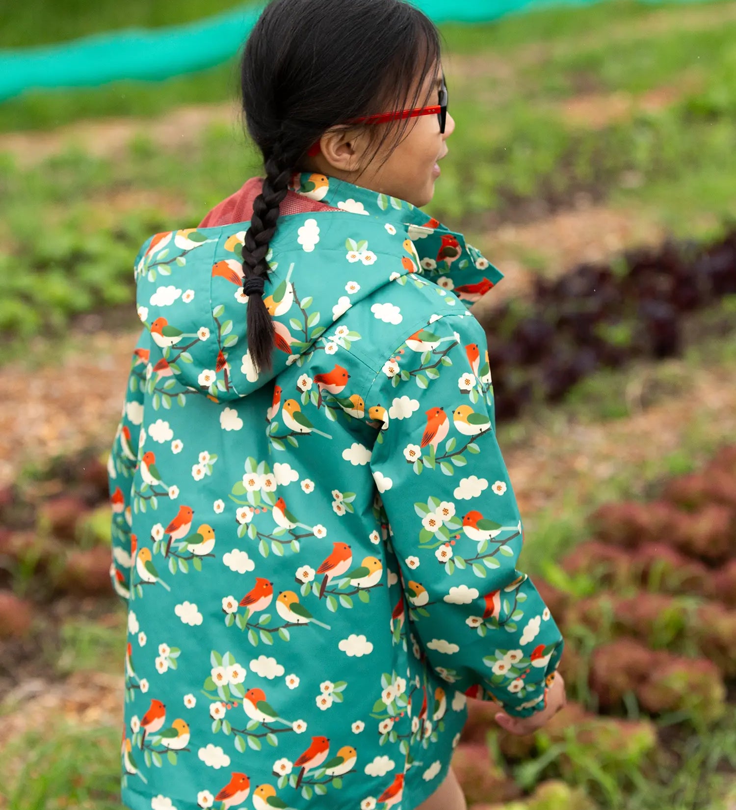 Back of a child wearing the Little Green Radicals turquoise birds recycled waterproof kids raincoat. Repeat pattern featuring bird on blossom branches.