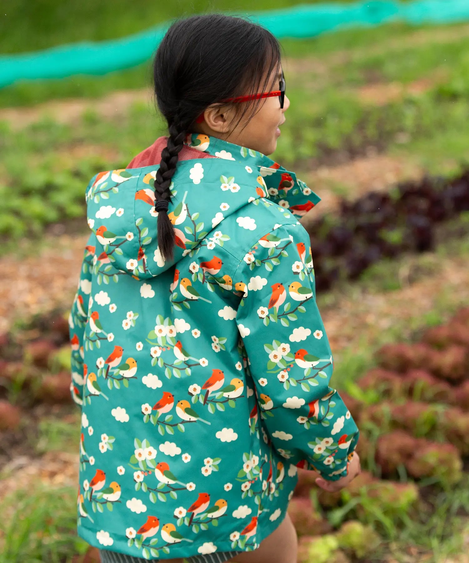 Back of a child wearing the Little Green Radicals turquoise birds recycled waterproof kids raincoat. Repeat pattern featuring bird on blossom branches.