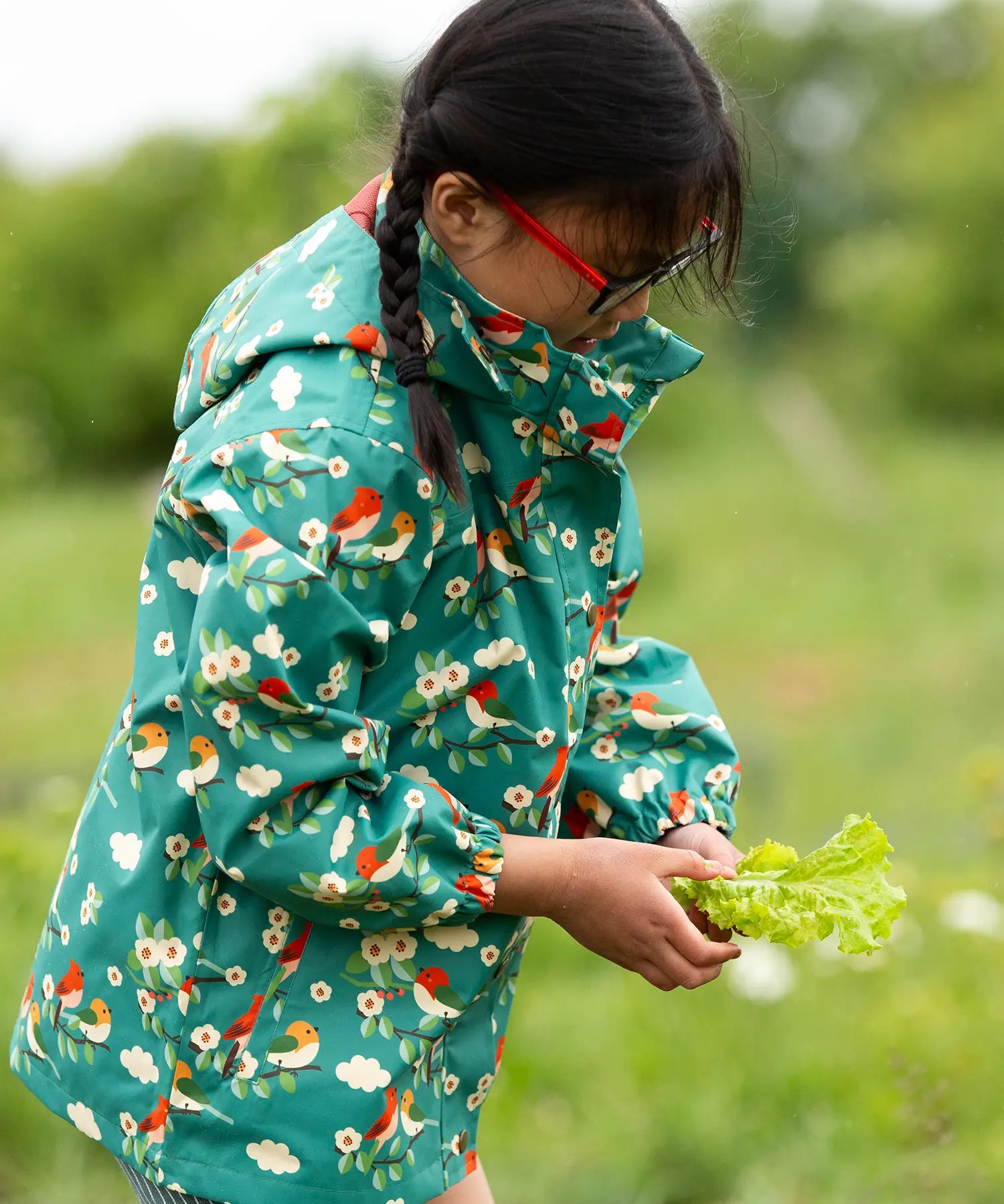 A child wearing the Little Green Radicals turquoise birds recycled waterproof kids raincoat. Repeat pattern featuring bird on blossom branches.