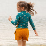 A child wearing the Little Green Radicals Teal Rainbows UPF 50+ Sunsafe Long Sleeve Rash Vest with yellow gold shorts. The child is standing by the sea facing away from the camera showing the back of the vest.
