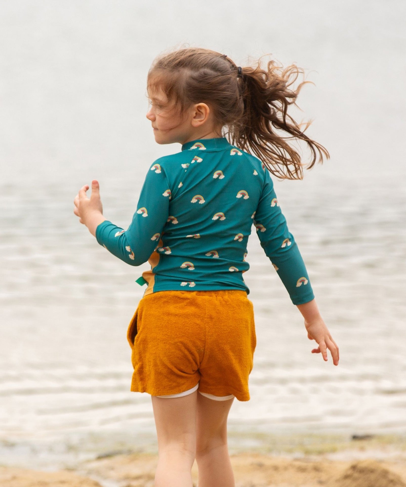 A child wearing the Little Green Radicals Teal Rainbows UPF 50+ Sunsafe Long Sleeve Rash Vest with yellow gold shorts. The child is standing by the sea facing away from the camera showing the back of the vest.