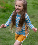 A child wearing the Little Green Radicals Children's Blue Whales design UPF 50+ Sunsafe Long Sleeve Rash Vest with yellow gold swim shorts.