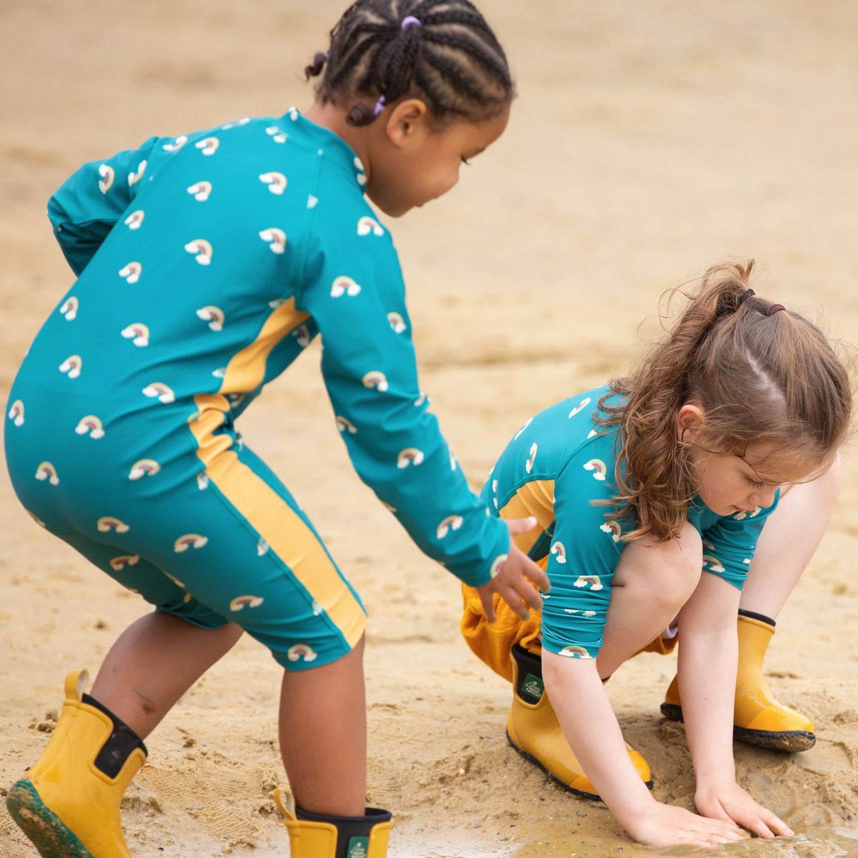 Two children plating on a sandy beach wearing teal rainbow swim wear from Little Green Radicals. 