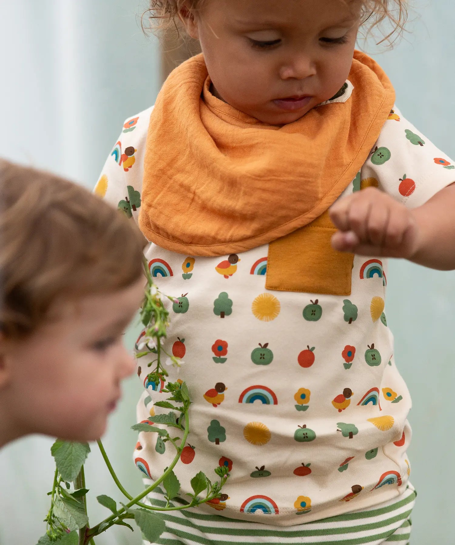 Close up of the repeat pattern print featuring nature themed illustrations top from the Little Green Radicals vintage style rainbows organic cotton t-shirt & jogger playset being worn by a toddler. 