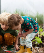 Close up of the Little Green Radicals weather knee patch blue and cream striped organic cotton joggers worn with a green apple print t-shirt and yellow wellington boots.