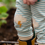 Close up of the Little Green Radicals weather knee patch blue and cream striped organic cotton joggers worn with a green apple print t-short and yellow wellington boots. The applique knee details features a white cloud and yellow lightning strike.