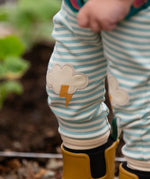 Close up of the Little Green Radicals weather knee patch blue and cream striped organic cotton joggers worn with a green apple print t-short and yellow wellington boots. The applique knee details features a white cloud and yellow lightning strike.