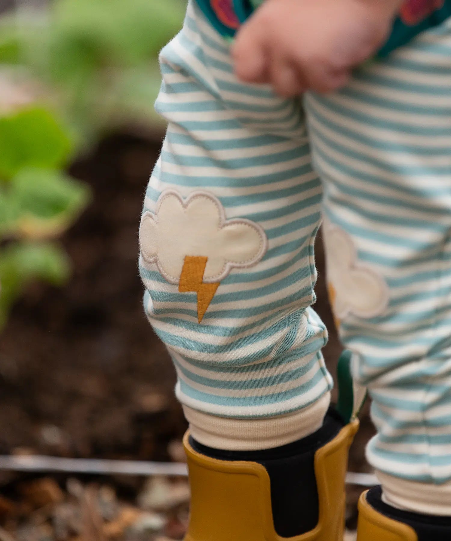 Close up of the Little Green Radicals weather knee patch blue and cream striped organic cotton joggers worn with a green apple print t-short and yellow wellington boots. The applique knee details features a white cloud and yellow lightning strike.