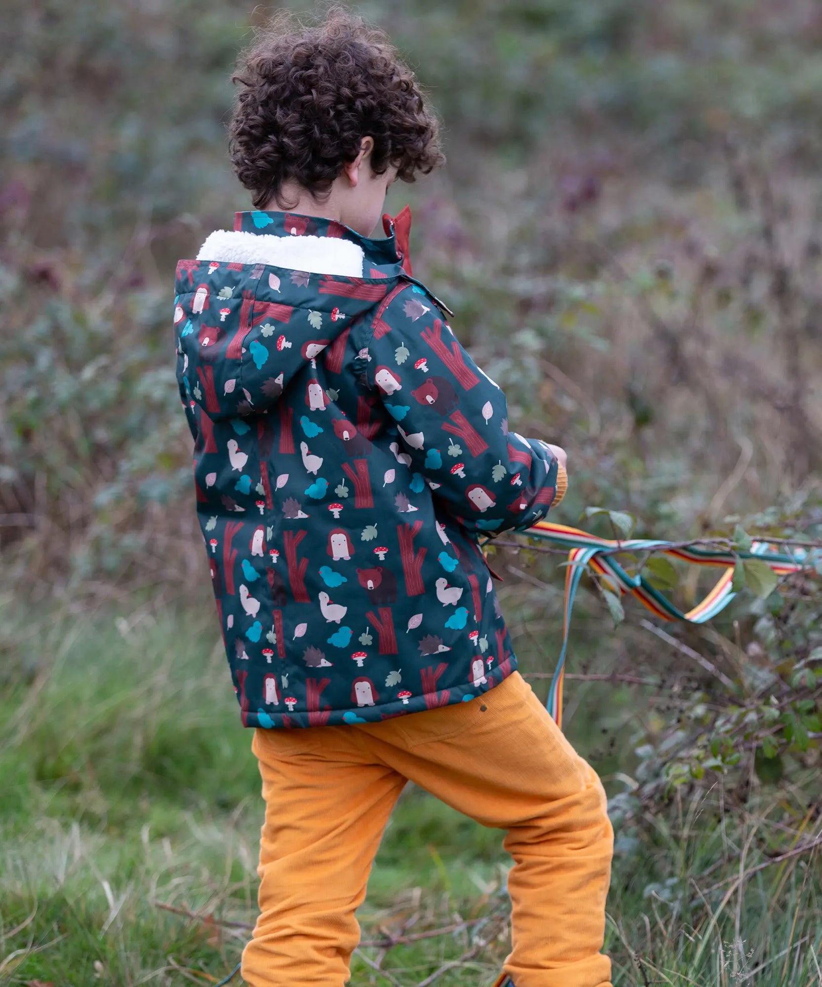 Child wearing a Little Green Radicals woodland world green recycled waterproof adventure winter coat with yellow gold trousers. View of the back showing the lined hood.
