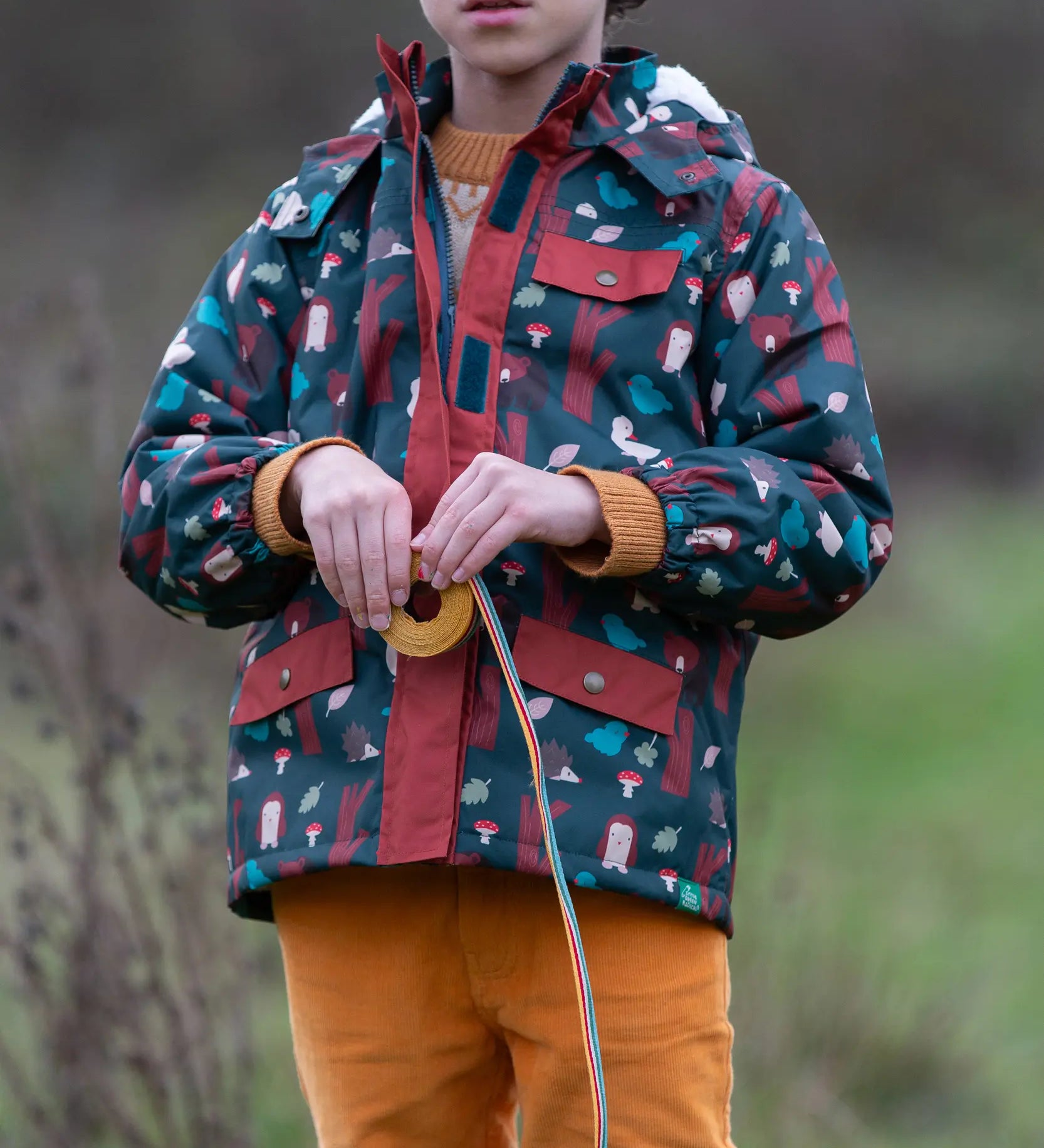 Child wearing a Little Green Radicals woodland world green recycled waterproof adventure winter coat with yellow gold trousers.