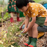 Child wearing the Little Green Radicals yellow gold summer pears short sleeve organic cotton t-shirt with green muslin shorts and green and red wellington boots. 