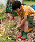 Child wearing the Little Green Radicals yellow gold summer pears short sleeve organic cotton t-shirt with green muslin shorts and green and red wellington boots. 