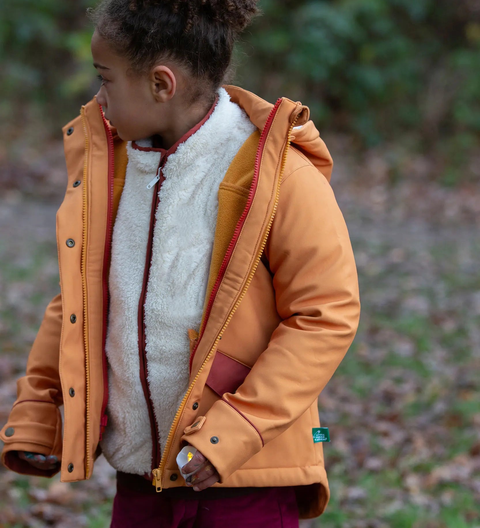 Child wearing a Little Green Radicals  yellow gold recycled waterproof 3 in 1 coat with berry purple trousers. The front zip is fully open.
