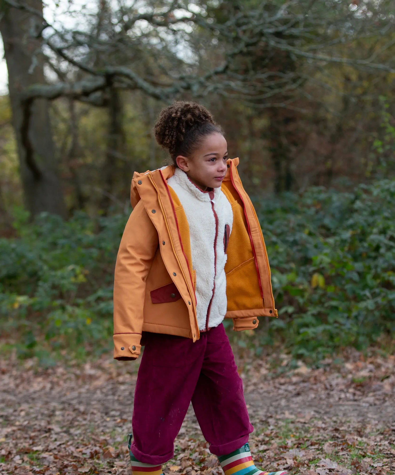 Child wearing a Little Green Radicals  yellow gold recycled waterproof 3 in 1 coat with berry purple trousers. The front zip is fully open.
