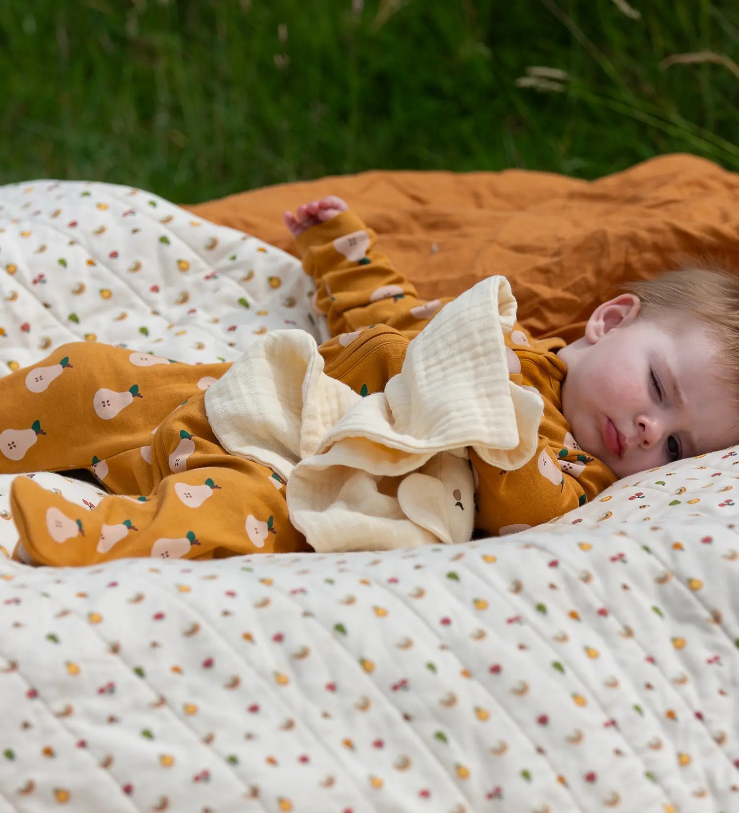 Little Green Radicals summer pears repeat print zip organic cotton footed babygrow on a baby laying on a patterned blanket. 