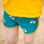 A close up of a child's legs wearing the Little Green Radicals Teal Rainbows UPF 50+ Recycled Swim Shorts.