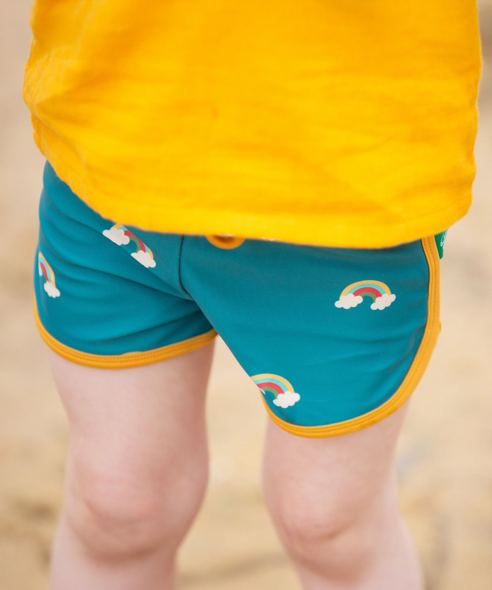 A close up of a child's legs wearing the Little Green Radicals Teal Rainbows UPF 50+ Recycled Swim Shorts.