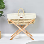 Little Green Sheep Moses Basket Stand pictured in a room with a grey coloured wall. A moses basket with a knitted insert is on the stand